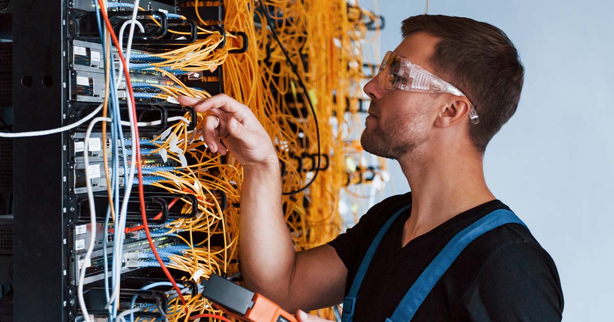 Young man in protective glasses works with internet equipment and wires in server room